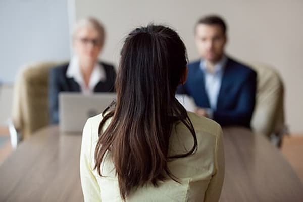 Woman sitting at conference table in front of two stern business people, being questioned during her interview